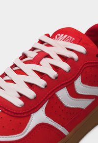 Κόκκινο αθλητικό παπούτσι με λευκές ζιγκ-ζαγκ ρίγες, λευκά κορδόνια και καφέ σόλα, με εμφανείς ραφές και μερικό κείμενο στη γλώσσα.