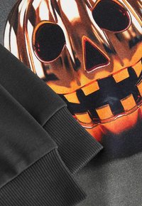 Svart sweatshirt med en orange grafik av ett jack-o'-lantern-ansikte med svarta accenter och ribbade ärmmuddar. Slät tygtextur.