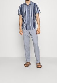 Camicia a maniche corte con righe verticali blu e bianche, con bottoni davanti, abbinata a pantaloni grigi chiari e sandali marroni.