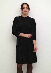 Svart klänning med vertikala ränder, hög hals, puffärmar och en avslappnad passform som faller ovanför knäet, tillverkad av textil med struktur.