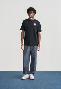 Μαύρο κοντομάνικο μπλουζάκι με πολύχρωμο λογότυπο στο στήθος, συνδυασμένο με χαλαρά γκρι παντελόνια και λευκά αθλητικά παπούτσια σε μπλε δάπεδο.