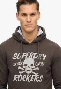 Sudadera con capucha en marrón oscuro con un estampado gráfico que presenta una calavera y huesos cruzados. Cordones y textura de tela suave.
