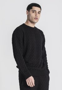 Suéter negro de punto con un patrón de trenzas texturizado, escote redondo y puños y dobladillo acanalados. Corte casual, material suave.