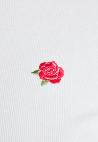 Biała, prążkowana tkanina z małym, czerwonym haftowanym różą i zielonymi liśćmi, z szczegółowym wzorem podkreślającym teksturę i kontrast kolorów.
