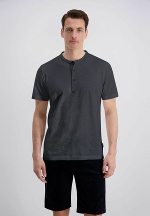 Man staat, draagt een donkergrijze Henley met korte mouwen en zwarte korte broek tegen een effen witte achtergrond.