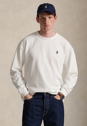 Νέος άνδρας με λευκό πουκάμισο μακρυμάνικο, σκούφο σκούρο μπλε και σκούρα τζιν, στέκεται με τα χέρια στις τσέπες μπροστά σε απλό φόντο.