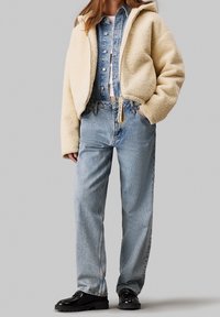 Kurtka z polarowym podbiciem w kolorze kremowym, krótka, założona na jasnoniebieską koszulę dżinsową, zestawiona z jasnoniebieskimi spodniami dżinsowymi o wysokim stanie i czarnymi błyszczącymi butami.