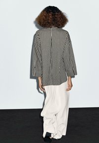 Black and white striped oversized shirt with loose sleeves, paired with cream wide-leg pants, viewed from the back against a pale background.