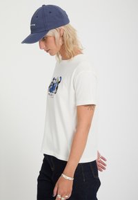 Ξανθό άτομο που φοράει λευκό γραφικό T-shirt, σκούρο παντελόνι, μπλε καπέλο, ασημένια κρίκους σκουλαρίκια και δαχτυλίδια, στραμμένο αριστερά με απλό φόντο.