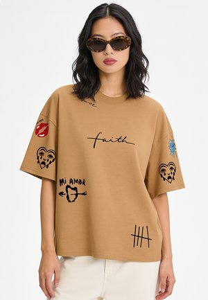 Femme aux cheveux noirs ondulés portant des lunettes de soleil écaille de tortue et un t-shirt beige oversize avec des imprimés graphiques noirs et rouges et un pantalon blanc.