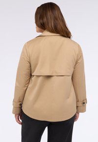 Beige lättviktsjacka med bakslå, knäppta ärmmuddar och skräddarsydd design. Den släta texturen och de enkla linjerna framhäver silhuetten.