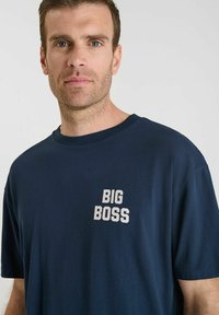 Man met kort bruin haar, gekleed in een marineblauwe T-shirt met "BIG BOSS" in witte letters op de borst, staande tegen een effen lichte achtergrond.