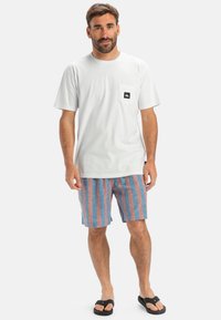 Bílé bavlněné tričko s černým logem na kapse, spárované s modro-růžovými pruhovanými šortkami a černými žabkami. Jednoduchý casual outfit.