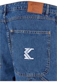 Jeans de mezclilla de color azul, con una etiqueta de cuero que dice "KARL KANI" y un logo bordado en el bolsillo trasero.