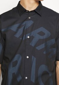 Černé tričko s krátkým rukávem se velkým tmavě modrým grafickým potiskem, klasickým límcem a knoflíkovým zapínáním vpředu s bílými knoflíky.