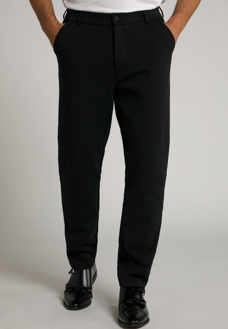 Hombre con pantalones negros a medida con bolsillos laterales y zapatos negros de cuero con doble hebilla monk en suelo gris claro.