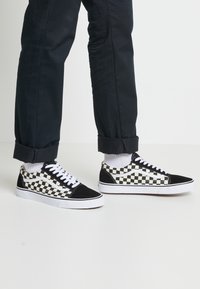 Baskets en toile à damier noir et blanc avec une semelle en caoutchouc, des lacets blancs et des accents noirs ; portées avec un pantalon foncé et des chaussettes blanches.