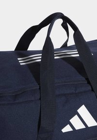 Námořnická sportovní taška s tlustými černými nosnými popruhy a bílým logem se třemi pruhy; má zipový vršek a texturovaný povrch látky.