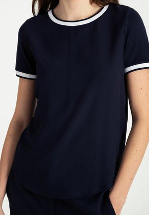 Kobieta ubrana w granatową koszulkę z krótkim rękawem, z białą lamówką na kołnierzyku i na zakończeniach rękawów, ręce częściowo w kieszeniach.