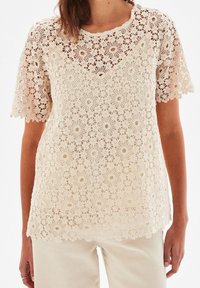 Haut en dentelle beige clair avec des motifs floraux et circulaires, encolure ronde et manches courtes. Design texturé, semi-transparent, matériau doux.