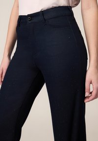 Pantaloni in denim blu scuro con sottili scintillii, dotati di vita aderente, tasche frontali e una texture liscia. Design classico a gamba dritta.
