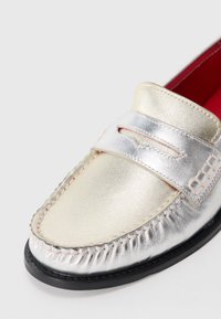 Metallic silver and gold loafers with stitched detailing, a broad strap, and a red interior. Flat sole with contrasting black edge.
