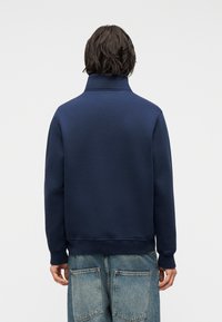 Άτομο με κοντά σκουρόχρωμα μαλλιά που φοράει μπλε σκούρο φούτερ με ψηλό γιακά και φαρδιά ανοιχτό μπλε τζιν, απεικονιζόμενο από πίσω.