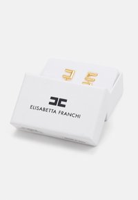 Balta Elisabethta Franchi dėžutė, dalinai atidaryta, rodo aukso geometrines auskarus viduje ant baltos pagalvėlės.