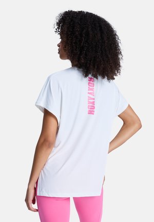 Kobieta o kręconych włosach, ubrana w białą koszulkę z pionowym różowym napisem na plecach oraz jaskrawo-różowe legginsy, stoi z jedną ręką na biodrze.