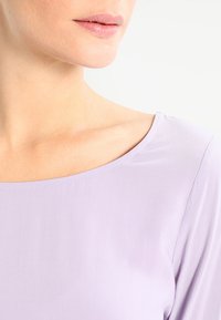Fliederfarbene Bluse mit weitem Ausschnitt und glattem Stoff. Sie hat einen subtilen Glanz und sitzt bequem auf den Schultern, mit minimalen Nahtdetails.