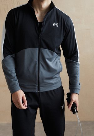 Mężczyzna w czarno-szarym dresie Under Armour trzyma skakankę na neutralnym tle.