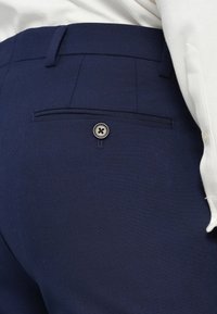 I pantaloni sartoriali blu navy presentano una tasca posteriore con chiusura a bottone nero. Il tessuto ha una trama liscia con un sottile motivo intrecciato.