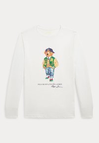 Bílé tričko s dlouhým rukávem s ilustrací medvěda v zelené varsity bundě, džínách a teniskách s barevnými akcenty.