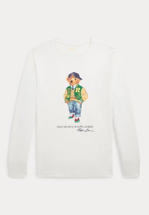 Witte T-shirt met lange mouwen met een illustratie van een beer in een groene varsityjack, spijkerbroek en sneakers met kleurige accenten.