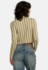 Mujer con cabello rizado oscuro, vistiendo una blusa cortada de mangas largas a rayas y jeans azul de tiro alto, de espaldas a la cámara.