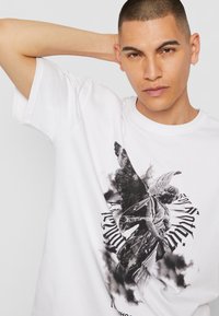 Hvid bomulds t-shirt med et sort grafisk tryk af en engel med vinger og tekst, sat mod en sky-lignende baggrund. Korte ærmer.