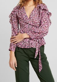 Blommig blus med rosa och vita blommor på mörk bakgrund. Har volangärmar, v-ringning och en knytning i midjan.