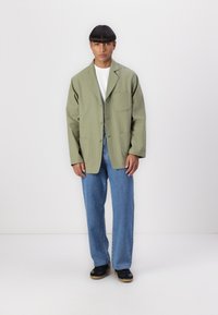 Light green oversized jacket with a single pocket, button closure, paired with light blue jeans and black shoes with a beige sole.