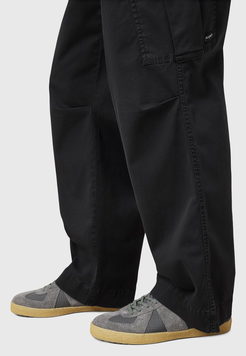 Pantaloni neri a gamba larga con una superficie morbida e tasche laterali, abbinati a sneakers grigie con una tomaia in suede e rete e suola in gomma.