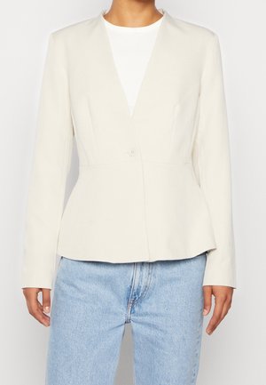 Persoon draagt een aansluitende gebroken witte blazer met één knoop over een wit topje en lichtblauwe spijkerbroek tegen een effen achtergrond.