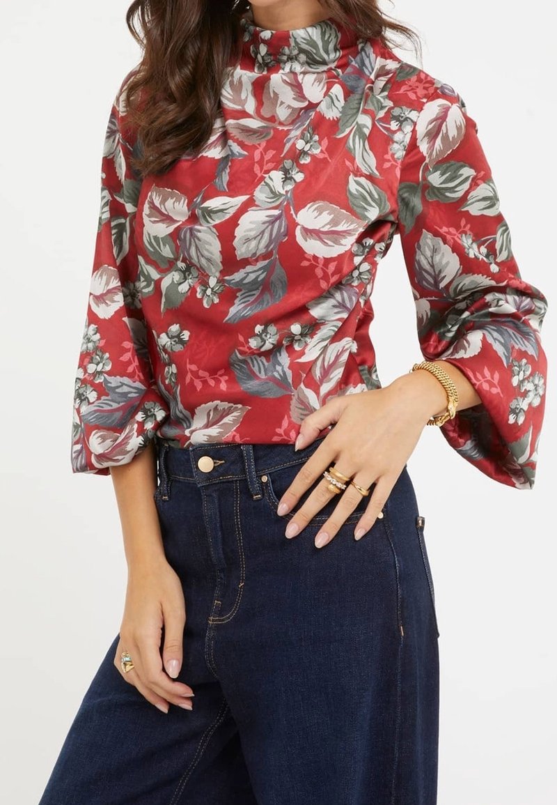 Rode bloementop met bell sleeves, versierd met groene bladeren en witte bloemen, gecombineerd met donkerblauwe high-waisted jeans. Gouden ringen om de hand.