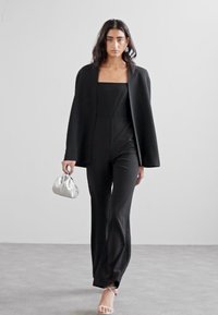 Tutina nera con scollatura quadrata, abbinata a un blazer nero strutturato, borsa metallica argento e sandali nudi con cinturini.