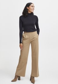 Un pull à col roulé noir associé à un pantalon large beige. Le pantalon présente des poches latérales et une texture lisse. Des escarpins pointus bruns complètent le look.