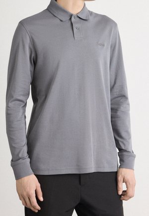 Modèle masculin portant un polo gris à manches longues avec un col et deux boutons, associé à un pantalon noir.