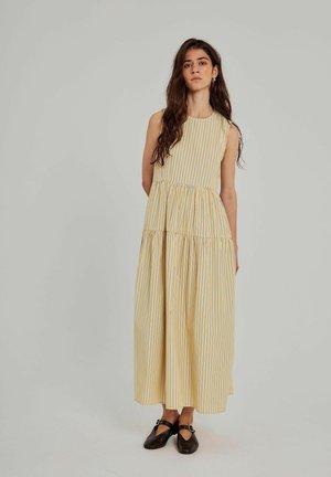 Long yellow dress with thin blue stripes, sleeveless, gathered waistline, flowing fabric. Paired with dark brown shoes.