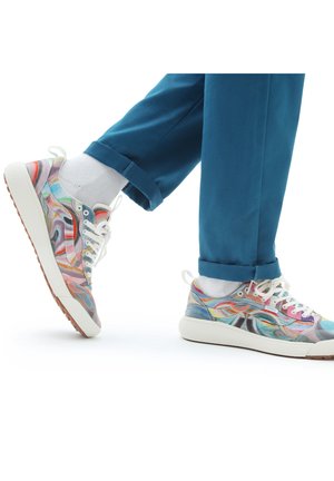 Personne portant un pantalon sarcelle et des baskets colorées à motifs, levant un pied comme pour faire un pas en avant sur un fond blanc.