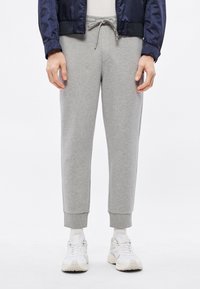 Szürke szabadidőnadrág bordázott mandzsettákkal és húzózsinóros derékrésszel, fehér tornacipővel párosítva. Az anyag puhanak tűnik, sima textúrával.