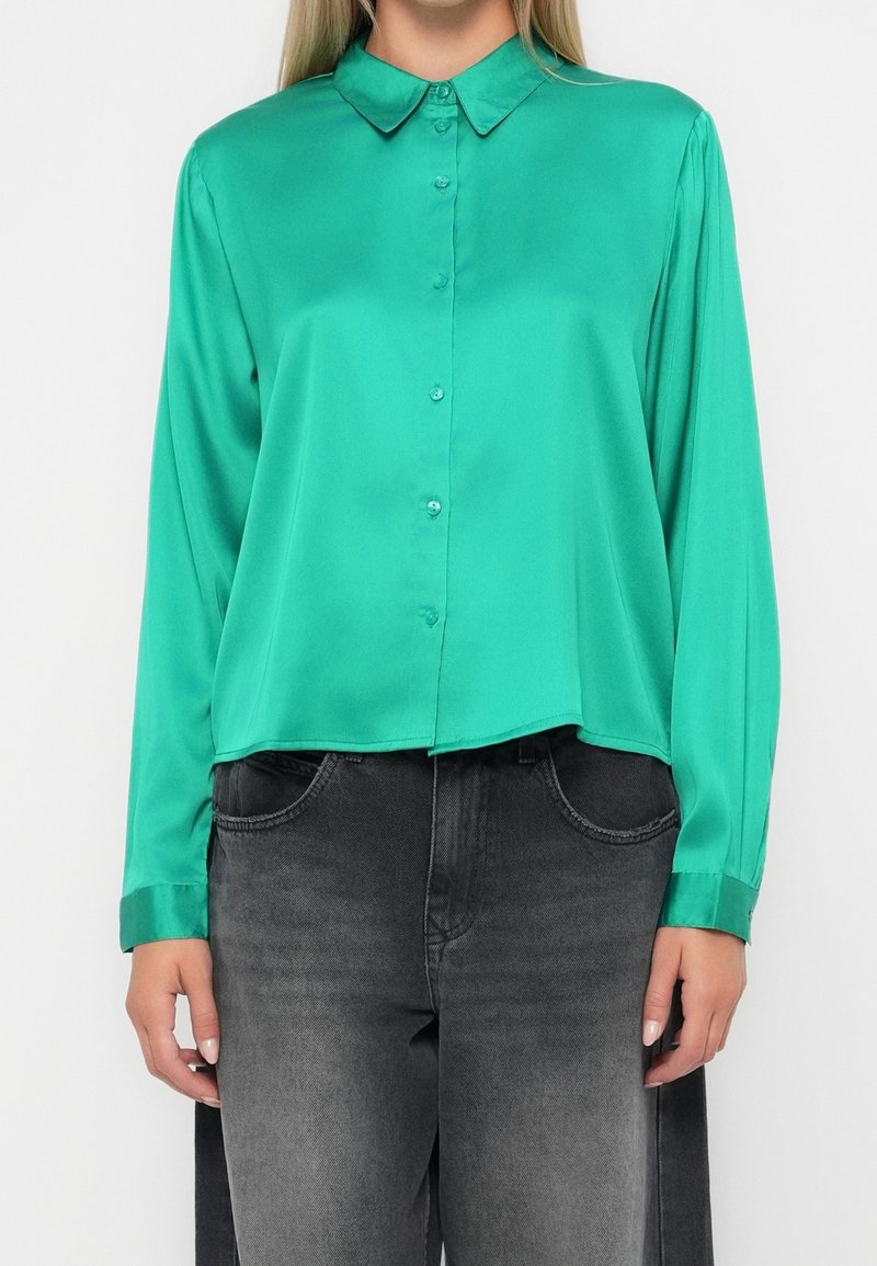 Femme portant une chemise verte à manches longues avec col et des jeans noirs délavés, debout devant un fond blanc uni.