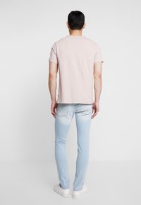 T-shirt rosa chiaro a maniche corte, in morbido cotone, dalla vestibilità rilassata. Indossata con jeans slim-fit blu chiaro e sneakers bianche, in piedi davanti a uno sfondo neutro.
