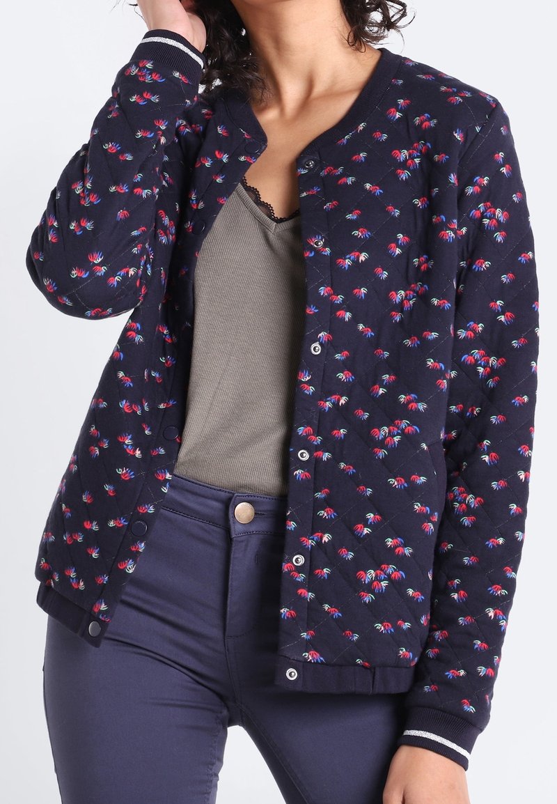 Femme portant une veste matelassée bleu marine avec de petits motifs floraux rouges et bleus, un débardeur côtelé gris et un pantalon bleu marine taille haute.